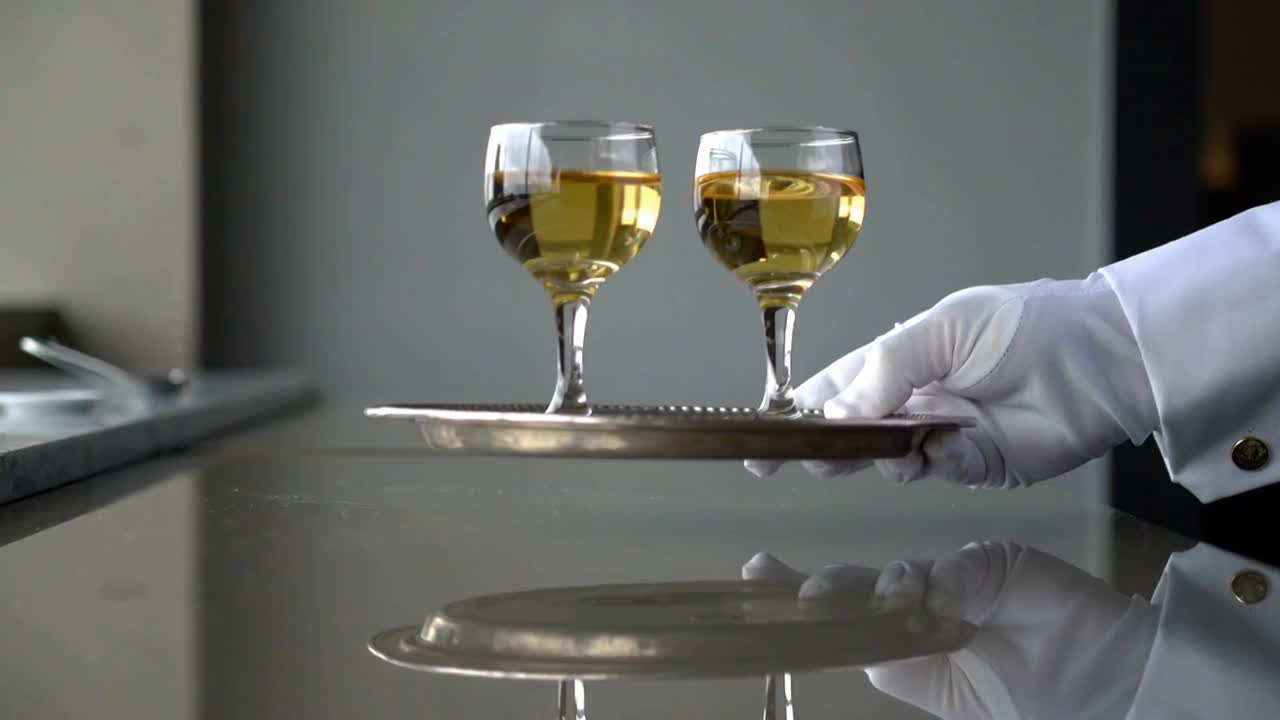 Lowering white-gloved hand placing silver tray with two pale wine glasses on shiny counter, serving