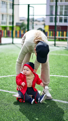 mom puts her little baby boy sitting on skate. adorable kid bends to touch the fake grass on the stadium. vertical video.