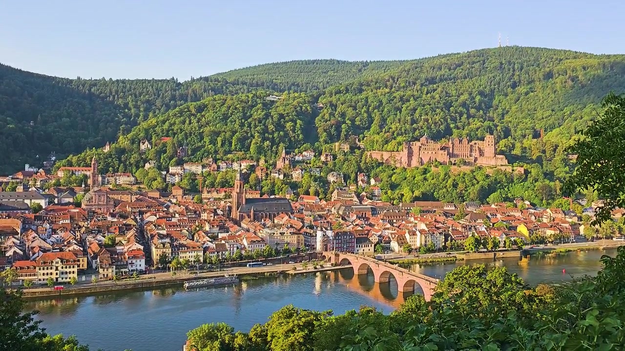 Vista elevata di Heidelberg, Germania, con rovine del castello sulla collina, il Ponte Vecchio che attraversa il fiume Neckar, tetti in tegole rosse, chiese storiche e colline boscose nella calda luce del sole serale