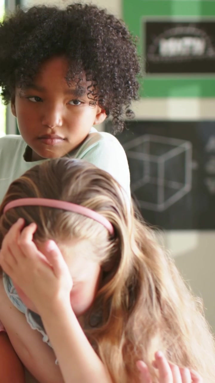 Video verticale: Reazione a un compagno che si copre il viso in classe, con un bambino dietro che si appoggia, rassicurante, con una maglietta chiara.