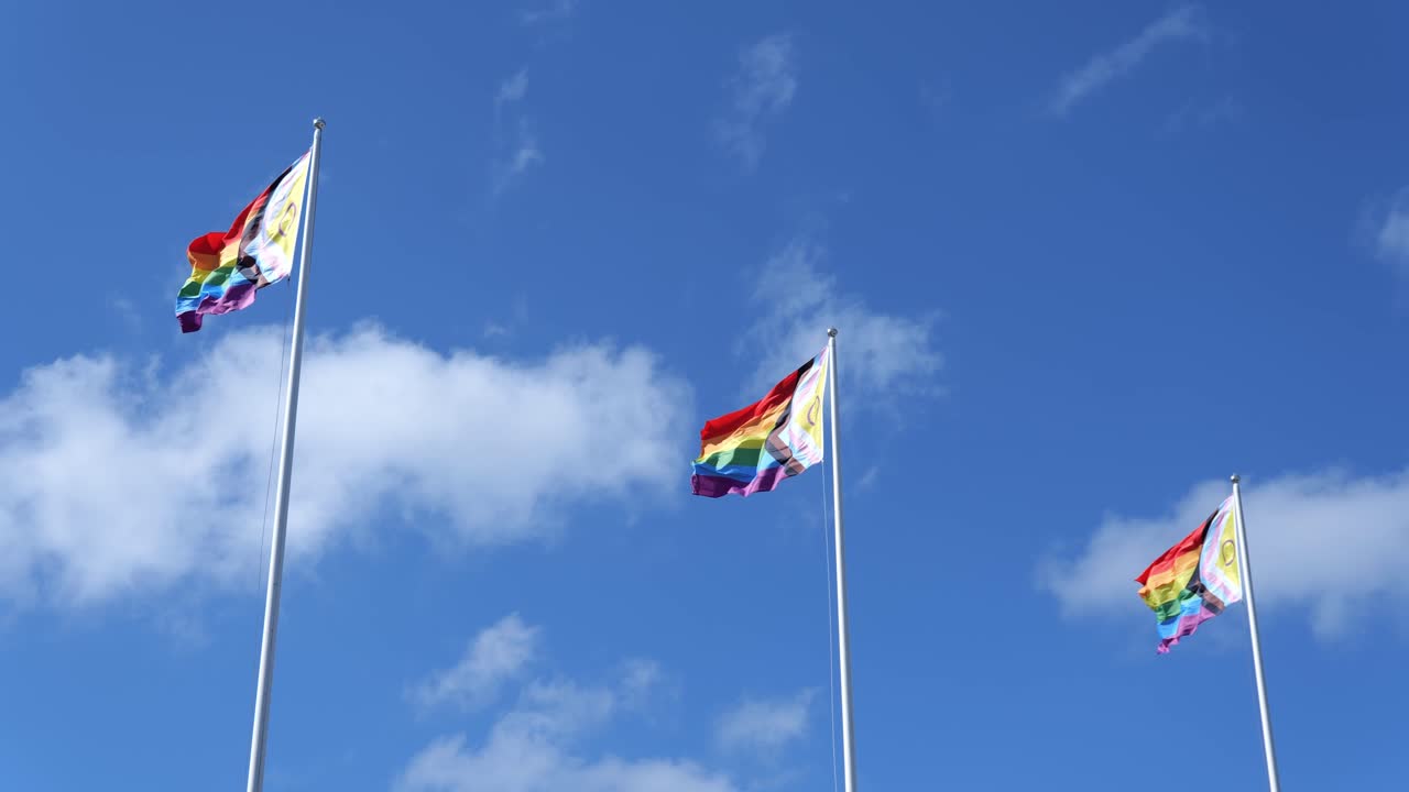 three rainbow pride flags waving proudly against a bright blue sky with fluffy clouds.