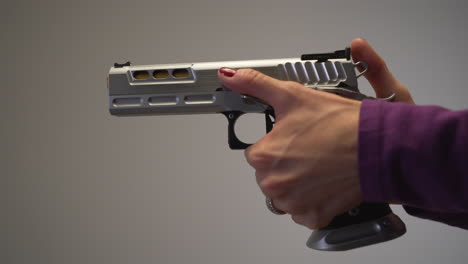 primer plano de mujer con uñas pintadas de rojo, amartillando y apretando el gatillo en una gran pistola plateada