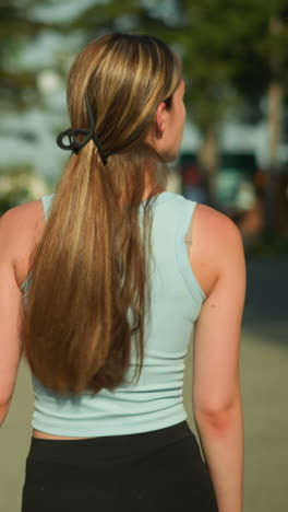 vista trasera de una joven con ropa atlética caminando por un camino entrelazado con el cabello atado hacia atrás, mirando de un lado a otro, la luz del sol mejora su movimiento mientras las personas están borrosas en el fondo