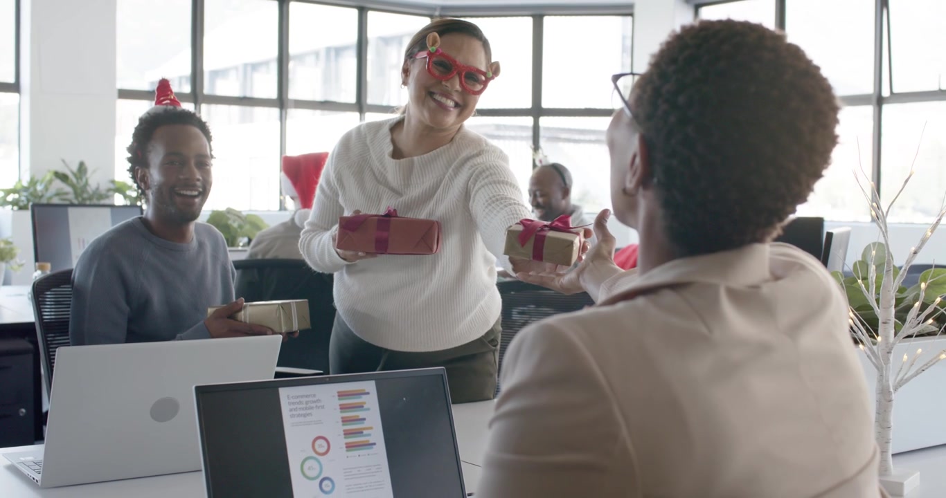 Diverse coworkers watching as woman wearing red glasses holding and offering gifts for Christmas