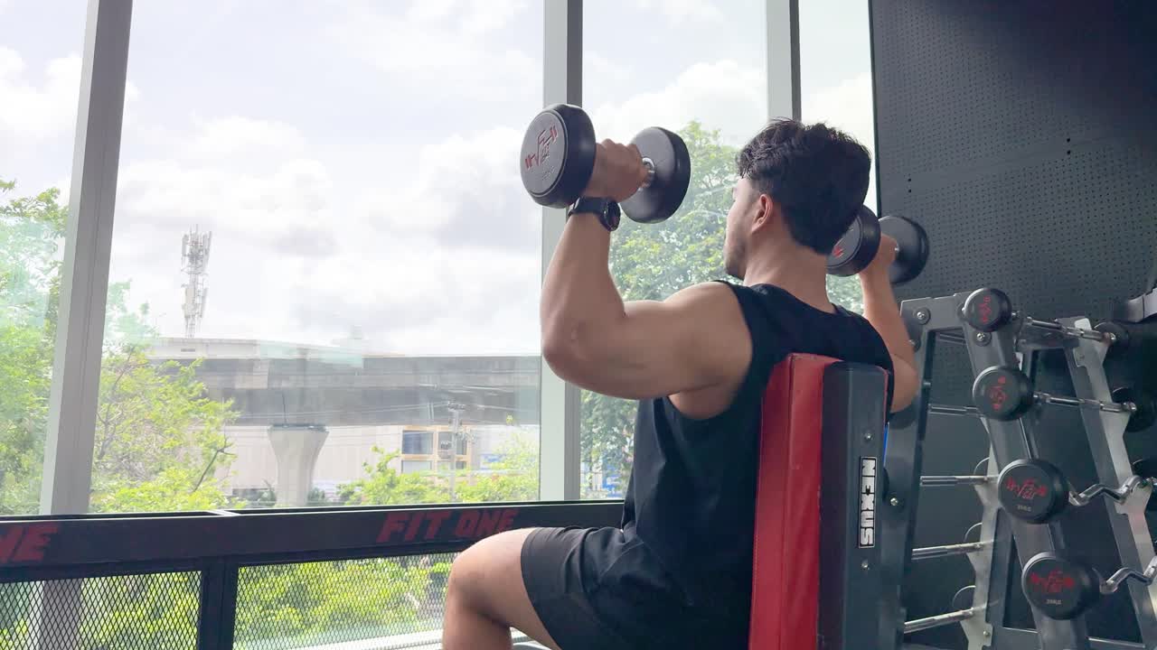 Athletic man lifts dumbbells overhead on bench in bright gym with large daylight windows