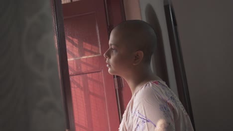 niña india calva mirando afuera desde la ventana en casa debido al bloqueo y cuarentena de covid-19