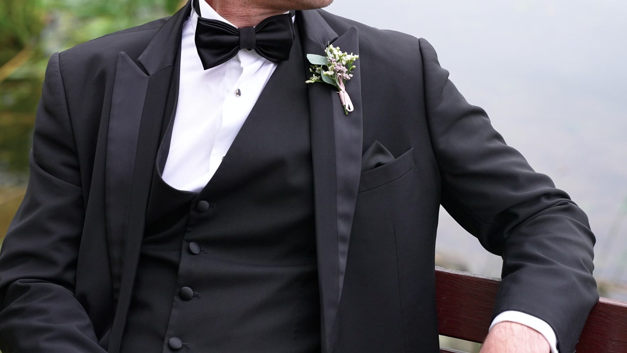Close-up shot of a well-dressed man in a black suit with a boutonniere, seated outdoors. The setting exudes sophistication with natural greenery and soft, ambient lighting.