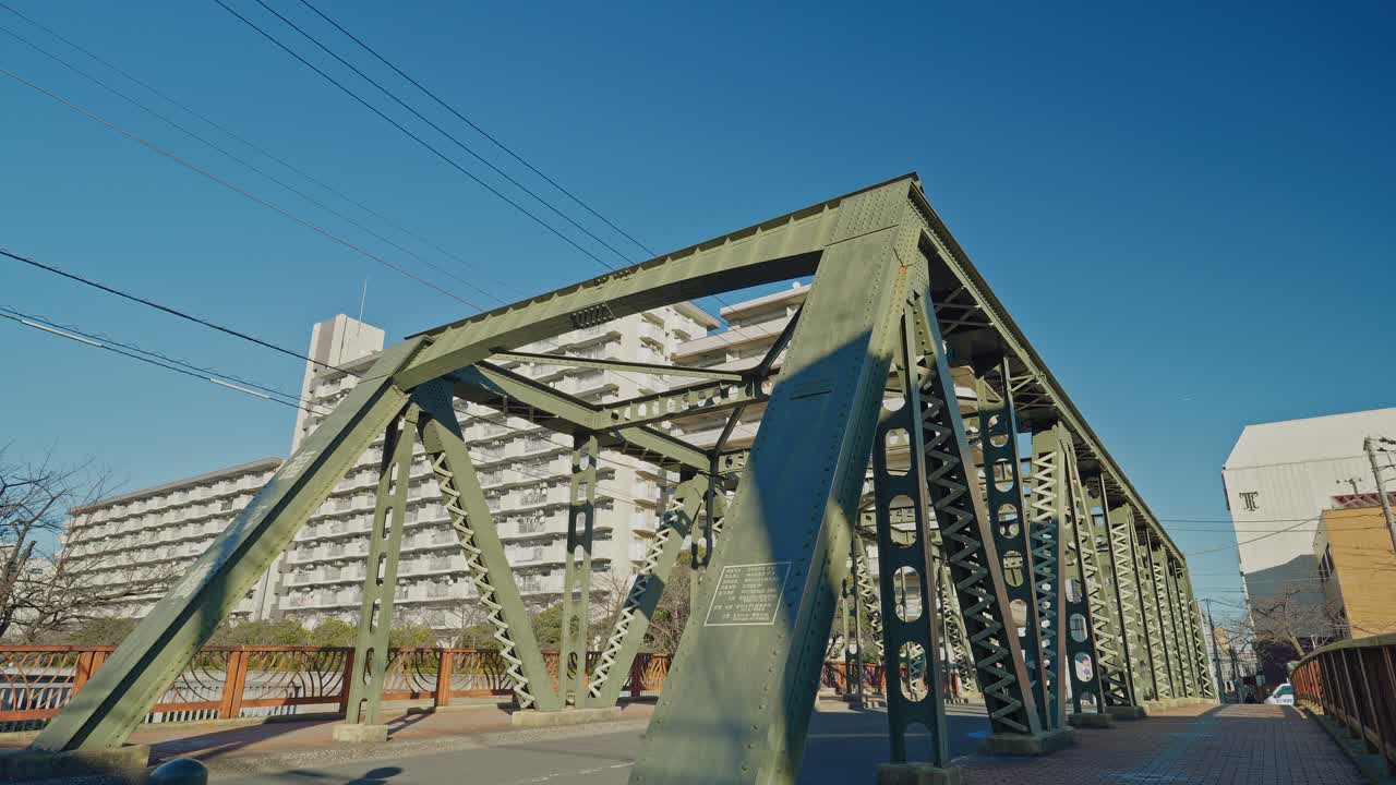 Fukuju Bridge's yellow-green steel truss viewed from ground level in Tokyo, with a tall residential tower rising behind the structure and bridge walkway visible, captured as a static wide shot in brig