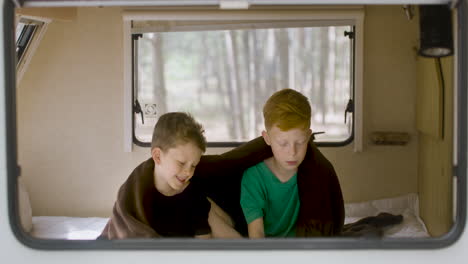 dos hermanos pequeños con una manta sobre los hombros sentados en la cama en una autocaravana y leyendo un libro