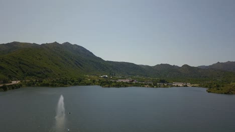 Vista-Aérea-Del-Lago-Con-Fuente-En-Verano-Con-Montañas-Verdes-Detrás-En-San-Luis,-Argentina