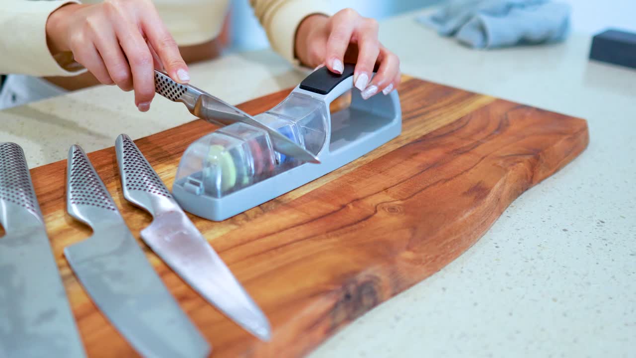 A person sharpens multiple kitchen knives using a manual whetstone sharpener on a wooden cutting board in a brightly lit kitchen
