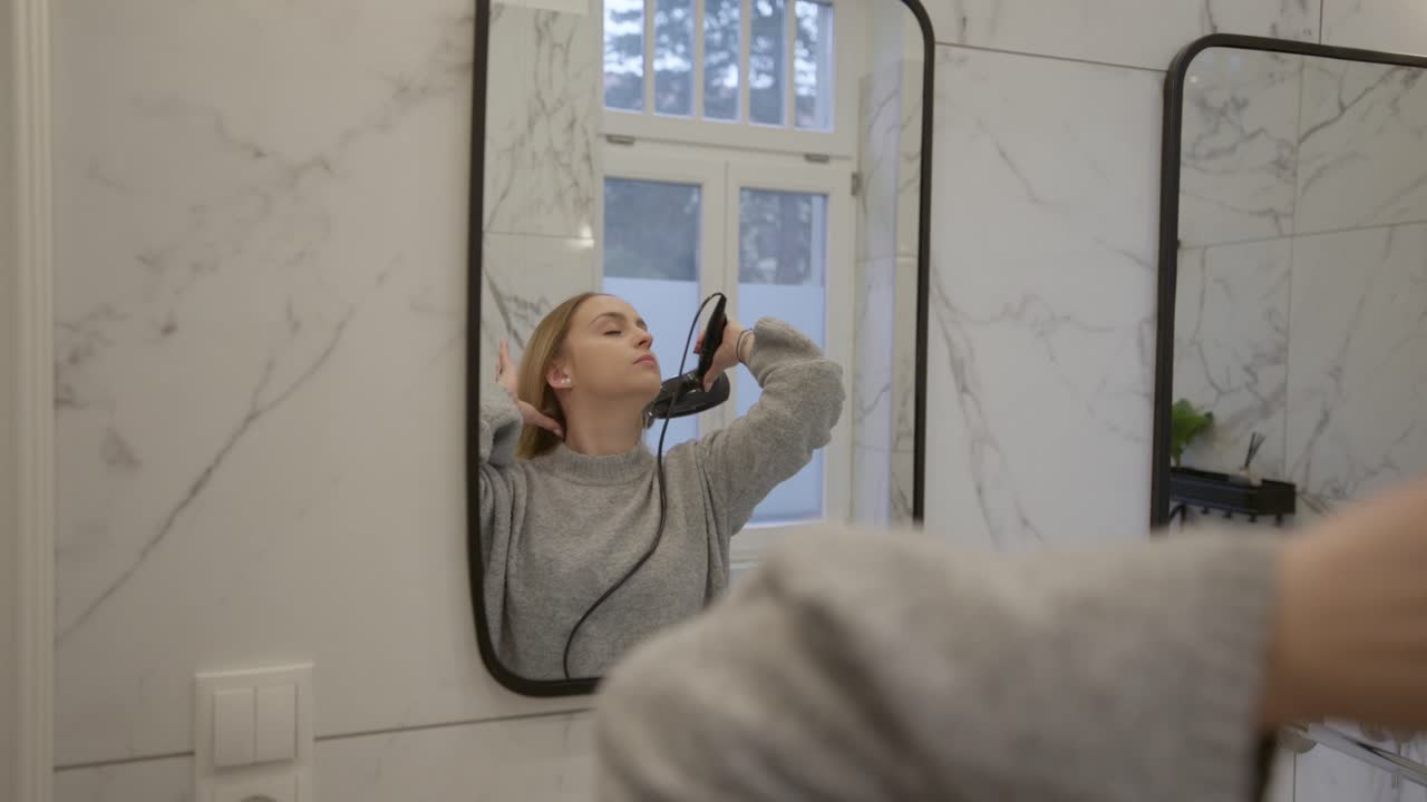 Una giovane donna bionda usa l'asciugacapelli mentre si acconcia i lunghi capelli e si guarda allo specchio in un bagno moderno
