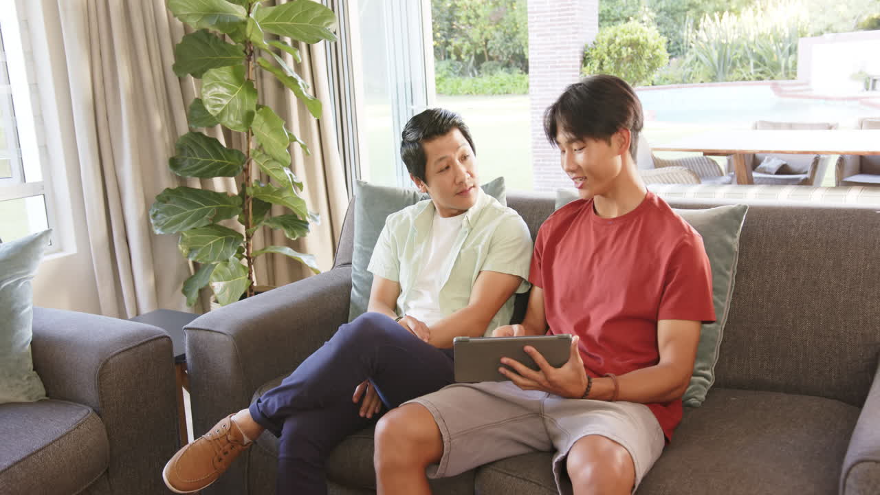 Sitting on couch, asian father and son discussing something on tablet in living room