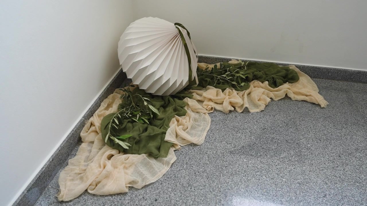 Una lampada di carta è posizionata accanto a un tessuto drappeggiato nei colori verde e beige su una superficie liscia. Questa composizione è ambientata in un semplice contesto interno che invita all'attenzione e alla concentrazione.