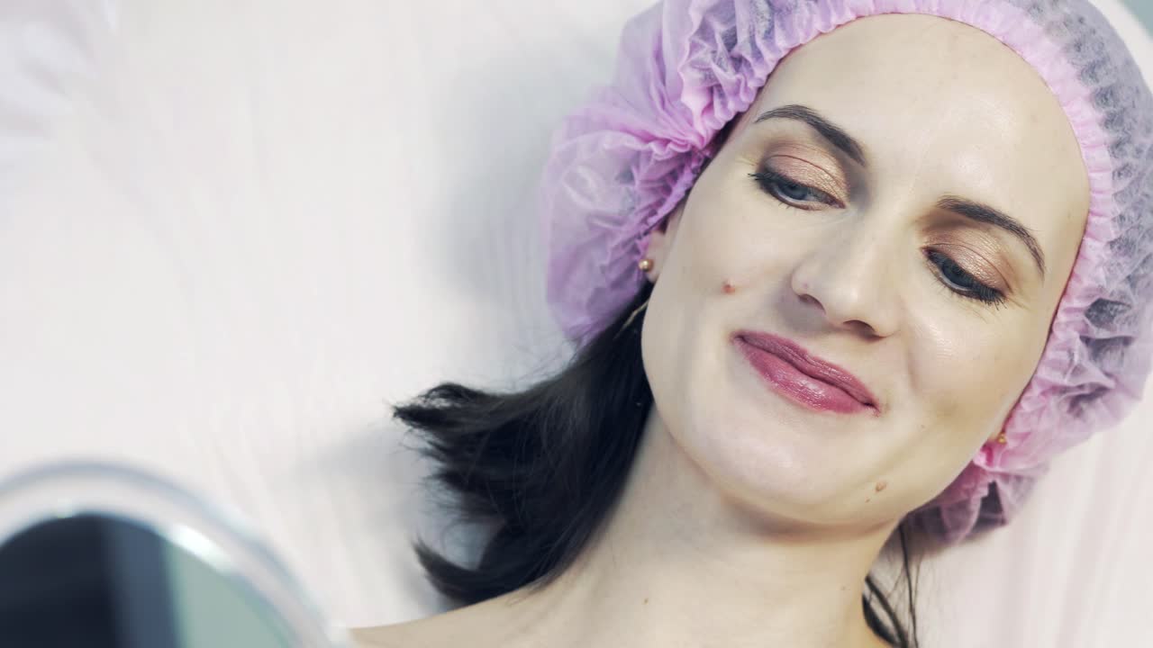 Patient is looking at herself in the mirror after the lip augmentation procedure and smiling in a beauty salon. Beautiful woman in medical cap and the mirror.