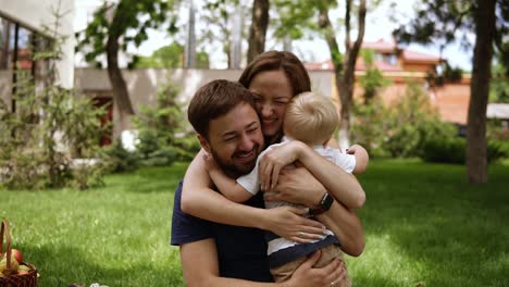 Happy-family-of-three.-Parents-in-casual-are-sitting-on-a-plaid-with-opened-hands-and-smiling-to-their-little-son.-Child-is-running-to-them.-Hugging,-smiling