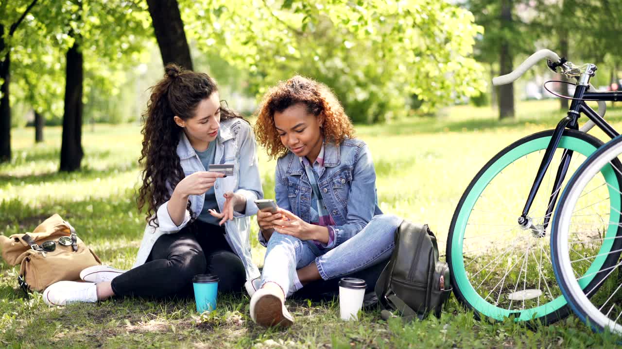dos mujeres jóvenes están pagando con tarjeta usando teléfono inteligente sentadas en el parque en la hierba sosteniendo tarjeta de crédito y teléfono luego haciendo high five y riendo. concepto de pago en línea.