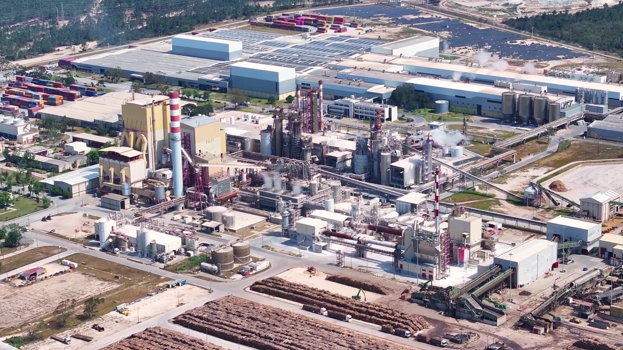 Expansive aerial view of a pulp and paper mill with its processing facilities, smokestacks, and large raw material yards for biomass energy