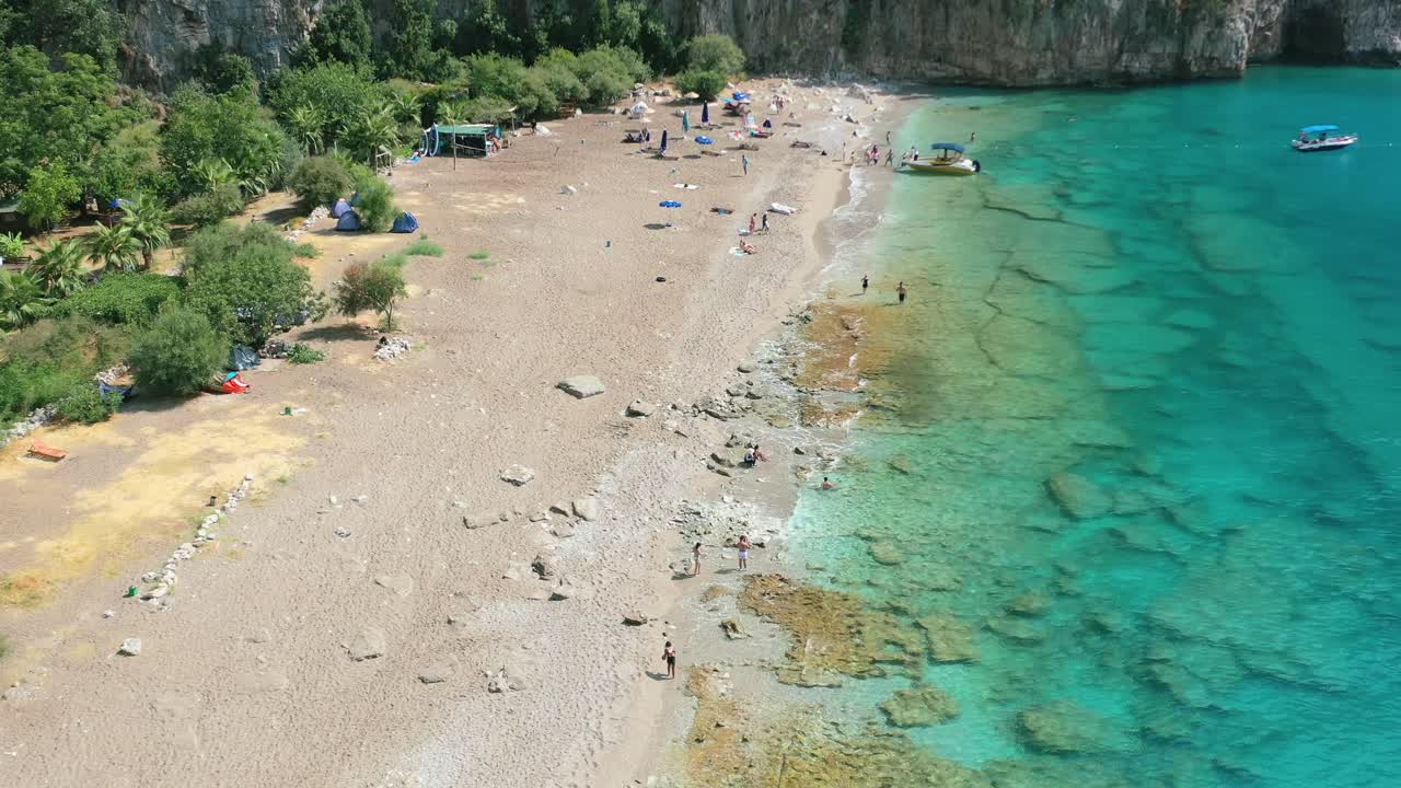 wide aerial of a rural coastal beach along the Mediterranean Sea known as Butterfly Valley in Fethiye Turkey as tourists walk alongside the turquoise blue tropical water on a sunny summer day