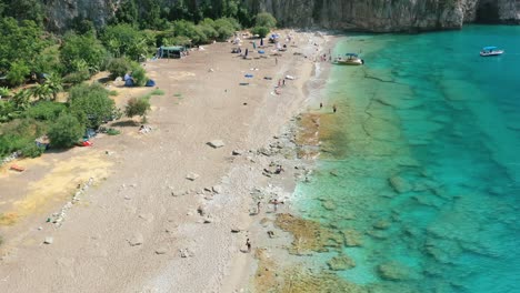 Breite-Luftaufnahme-Eines-Ländlichen-Küstenstrandes-Am-Mittelmeer,-Bekannt-Als-Schmetterlingstal-In-Fethiye,-Türkei,-Während-Touristen-An-Einem-Sonnigen-Sommertag-Am-Türkisblauen-Tropischen-Wasser-Entlang-Spazieren