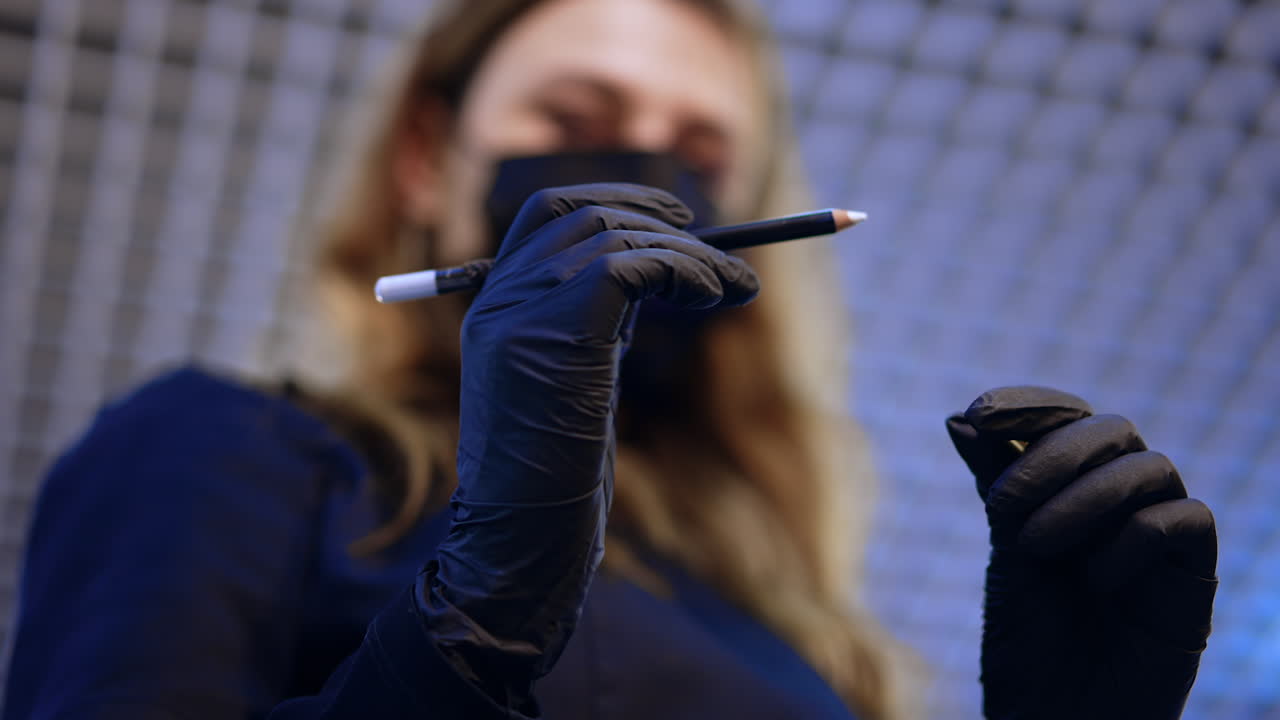 Unrecognized beauty master opens white pencil. Specialist in mask and gloves is going to line the brows for future permanent make up. Low angle view. Blurred backdrop.
