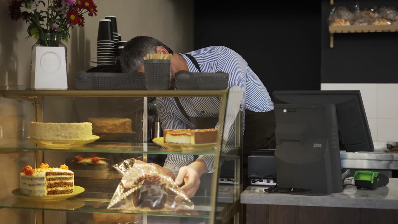 Young handsome man vendor in the apron bringing fresh cake to the counter in the bakery shot. Indoors