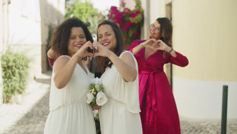 Afro-American-lesbian-couple-making-heart-with-their-hands,-kissing-and-posing-for-camera-with-friends-after-wedding-ceremony