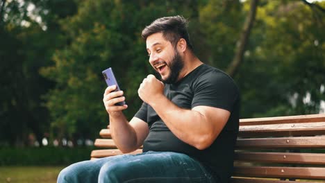 Young-man-celebrating-good-news-recieved-on-his-cell-phone-while-seated-at-a-park-bench