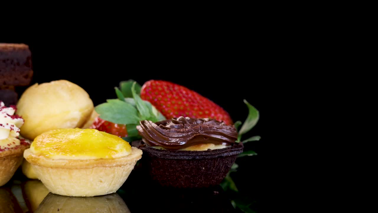 An array of brownies, cream puffs, tarts, and strawberries rotates on a glossy black surface under bright studio lighting, highlighting vibrant colors and textures