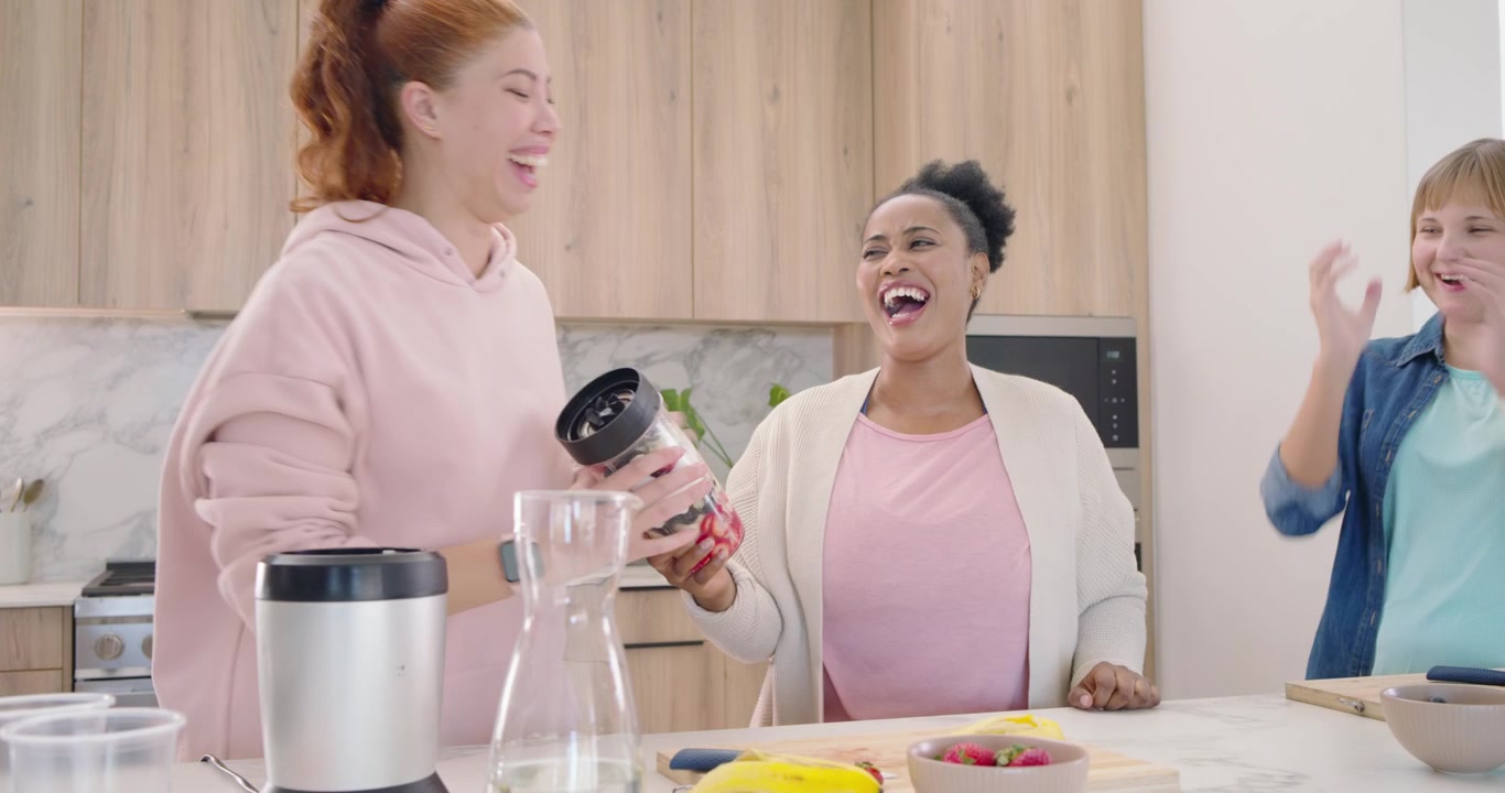 Amiche di diverse etnie che preparano il frullatore sull'isola della cucina, chiudendo il coperchio, avviando il frullatore e ridendo.