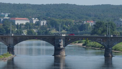 a red bus travels across a multi-arched stone bridge spanning a calm river, with lush green hills and urban buildings in the background under daylight