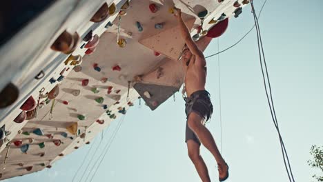 Bottom-view:-an-athletic-guy-in-black-shorts-with-a-sculpted-torso-climbs-up-the-climbing-wall-and-hangs-on-the-ledge-of-the-training-route-of-the-climbing-wall-on-a-sunny-summer-day