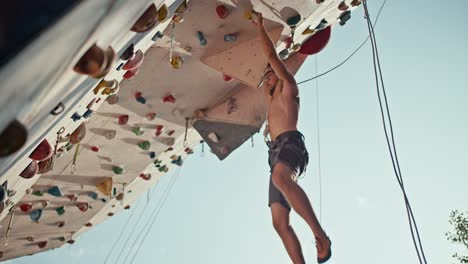 vista de abajo: un tipo atlético en pantalones cortos negros con un torso esculpido sube a la pared de escalada y se cuelga en la repisa de la ruta de entrenamiento de la parede de escalada en un soleado día de verano