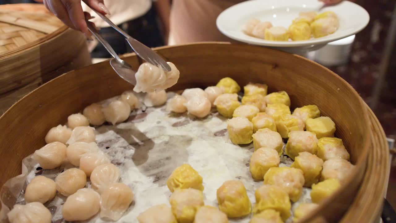 A person uses metal tongs to select shrimp dumplings and shumai from a bamboo steamer