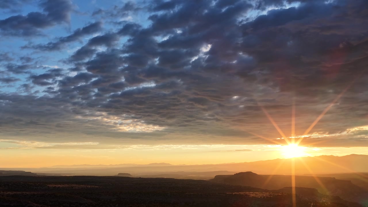 4K drone timelapse of a vibrant sunrise over the White Rock, New Mexico landscape. A dramatic blanket of shifting clouds illuminates with golden dawn colors above the vast, rugged desert terrain.