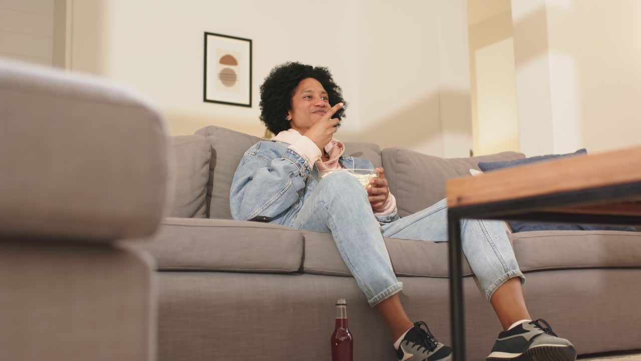 Uomo afroamericano di mezza età che guarda un film, preme il telecomando a casa, mangia popcorn e mostra stupore
