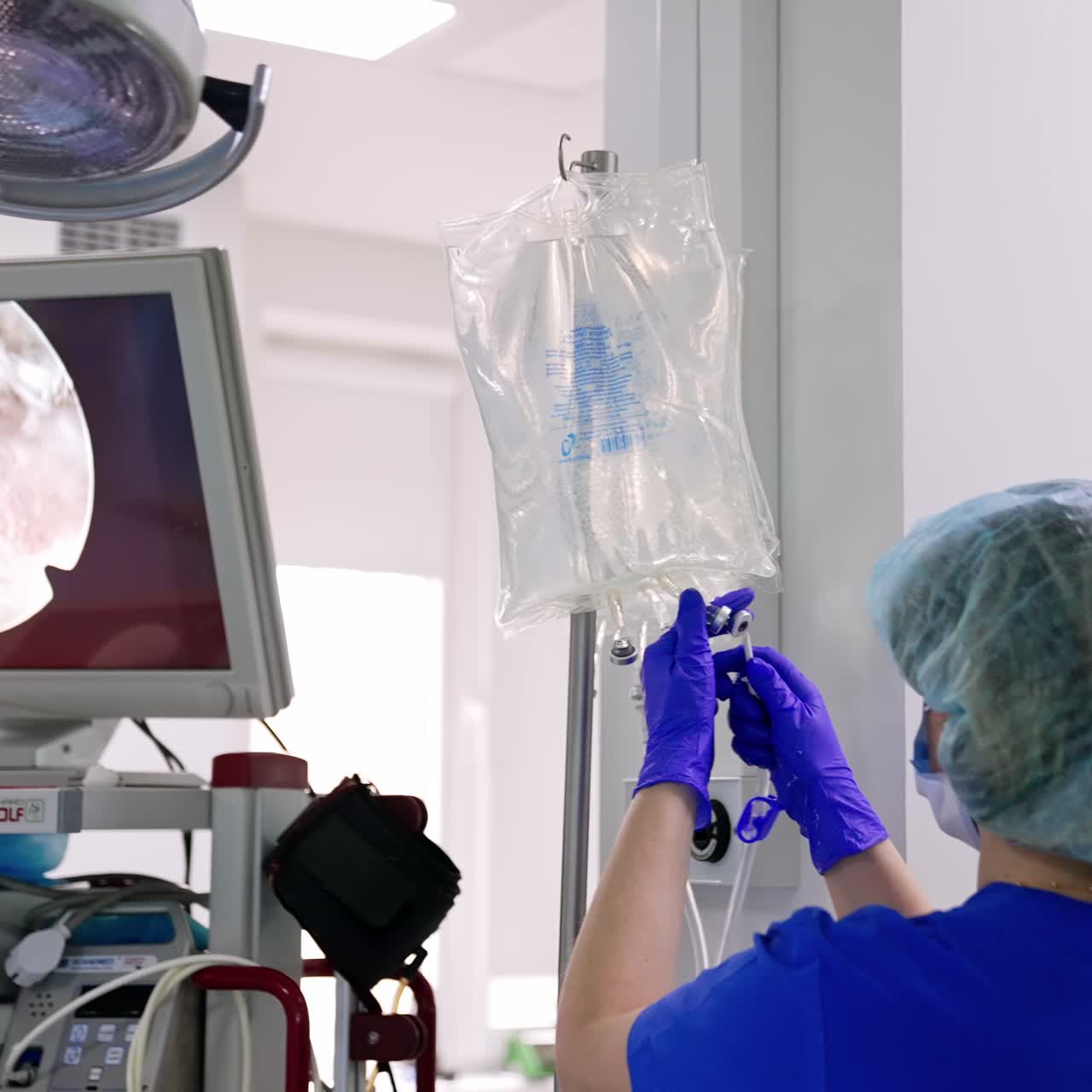 Vista posteriore di un'infermiera che sistema il conta gocce in sala operatoria. Un grande schermo accanto mostra l'operazione dall'interno