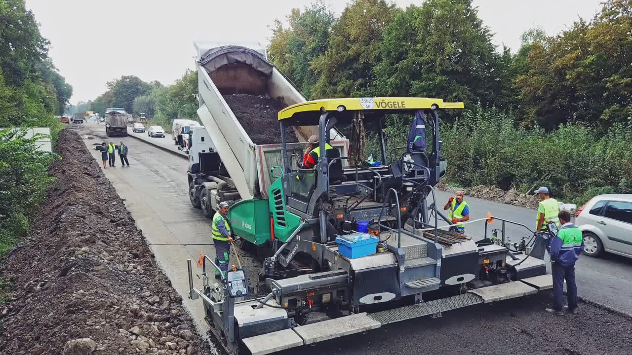 A truck tosses over bitumen into a wide paver to lay a new roadway surrounded of repairmen. Road construction equipment. Aerial view.