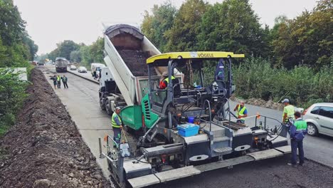 a truck tosses over bitumen into a wide paver to lay a new roadway surrounded of repairmen. road construction equipment. aerial view.