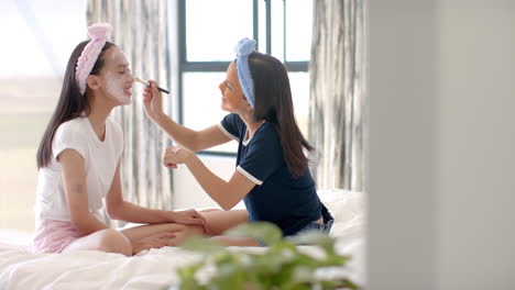 two biracial teenage girl friends sitting on bed at home, one applying facial mask to other
