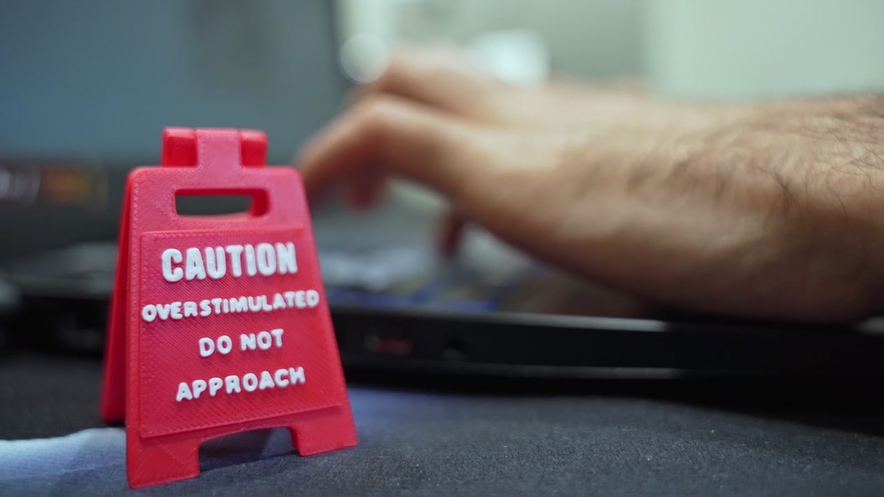 Close up of a red miniature caution sign reading overstimulated do not approach on a desk in front of a laptop. Concept of workplace stress, mental health, and neurodiversity at work.