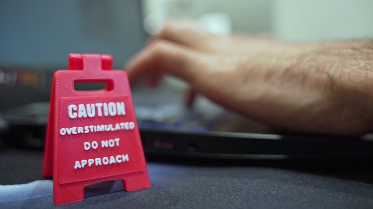 Close up of a red miniature caution sign reading overstimulated do not approach on a desk in front of a laptop. Concept of workplace stress, mental health, and neurodiversity at work.