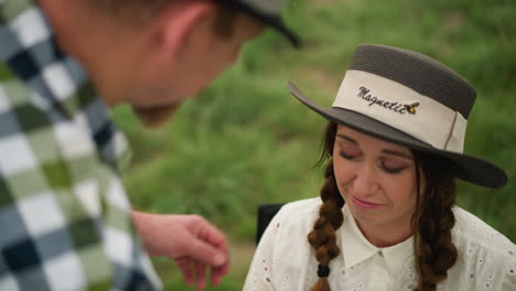 an artist in a checked shirt adjusts the face of a smiling woman with braided hair, wearing a stylish black hat and white dress, seated in a lush green field