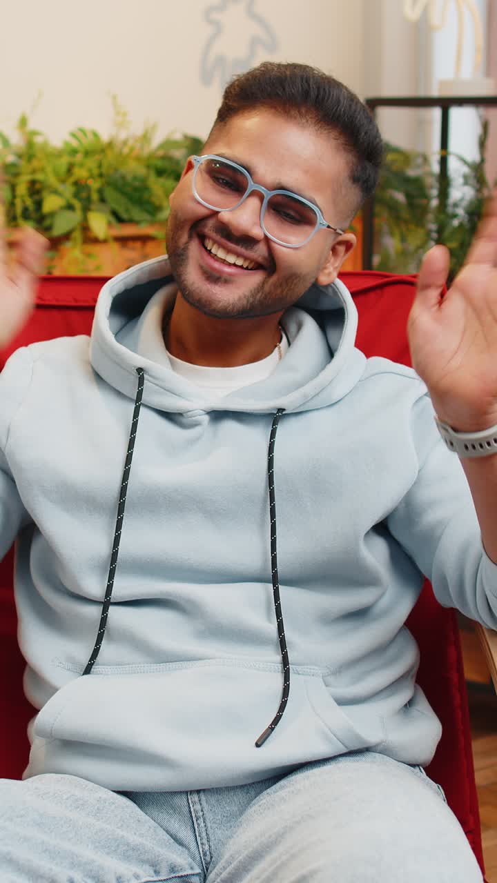 Indian man smiling friendly at camera waving hands gesturing hello hi greeting at home on chair