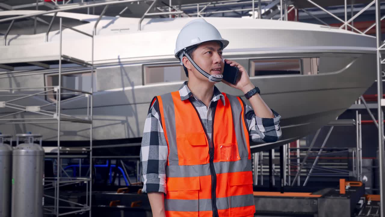 Ingeniero asiático con casco de seguridad hablando por teléfono mientras está de pie junto a la construcción de un yate en un astillero moderno
