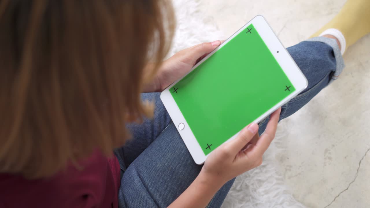 mujer joven asiática usando un dispositivo de tableta negra con pantalla verde. mujer asiática sosteniendo una tableta, desplazando páginas mientras está sentada en el sofá en la sala de estar. clave de croma.