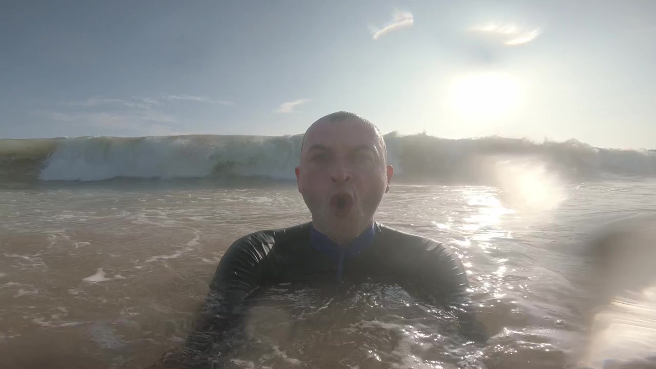Un homme fait face à la caméra au niveau de l'eau dans des vagues jusqu'à la poitrine alors qu'une vague océanique rétroéclairée se forme derrière lui, s'écrase sur sa tête et submerge brièvement la vue à Mirissa Beach, Sri Lanka