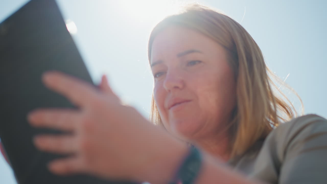 Close up of young woman admiring something on phone screen outdoors as sunlight creates warm lens flare around her face, capturing emotion, curiosity, and connection with digital world