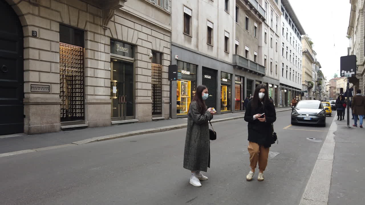 Slow-motion shot of two girls with masks in via Montenapoleone taking a picture to a pharmaceutical product in via Monte Napoleone, Milan, Italy in the late afternoon during a grey day.