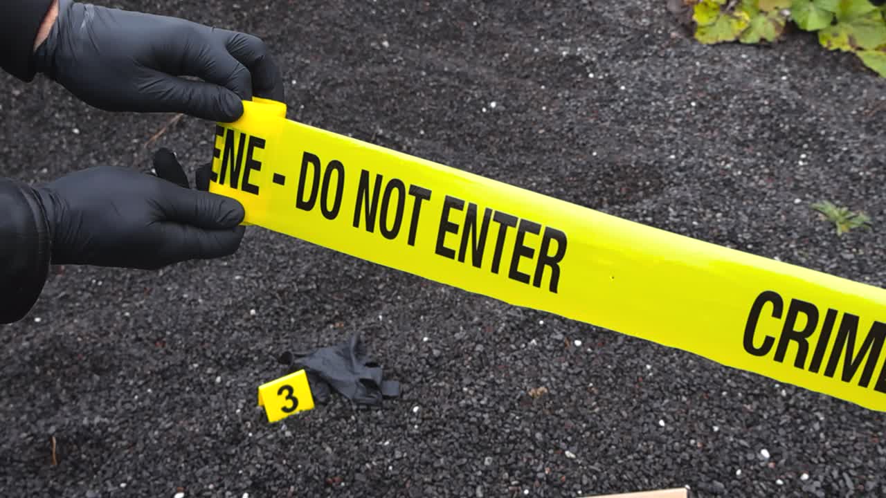 Police investigator or officer with black nylon gloves unrolling a crime scene yellow forensic investigation tape in front of a crime scene with a bloody knife and a glove as evidence, crime markers